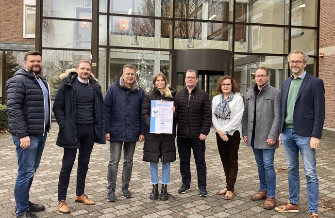 Eine Gruppe von Klinikvertreter:innen stehen vor dem Klinikeingang und halten das EMAS-Zertifikat in die Kamera.