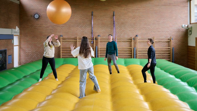 Personen stehen auf einem Luftkissen und werfen sich einen orangenen Ball zu.