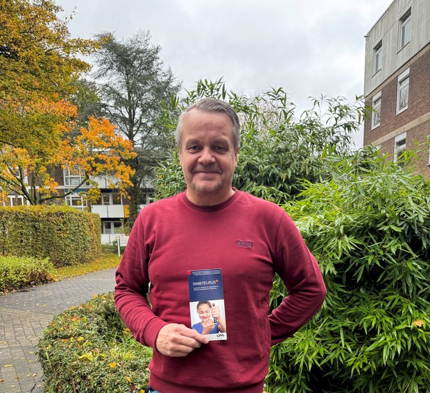 Ein Kollege lächelt in die Kamer und hält einen Flyer in der Hand