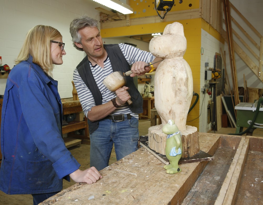 Zwei Menschen blicken auf eine Holzskulptur, diese wird von einer Person bearbeitet