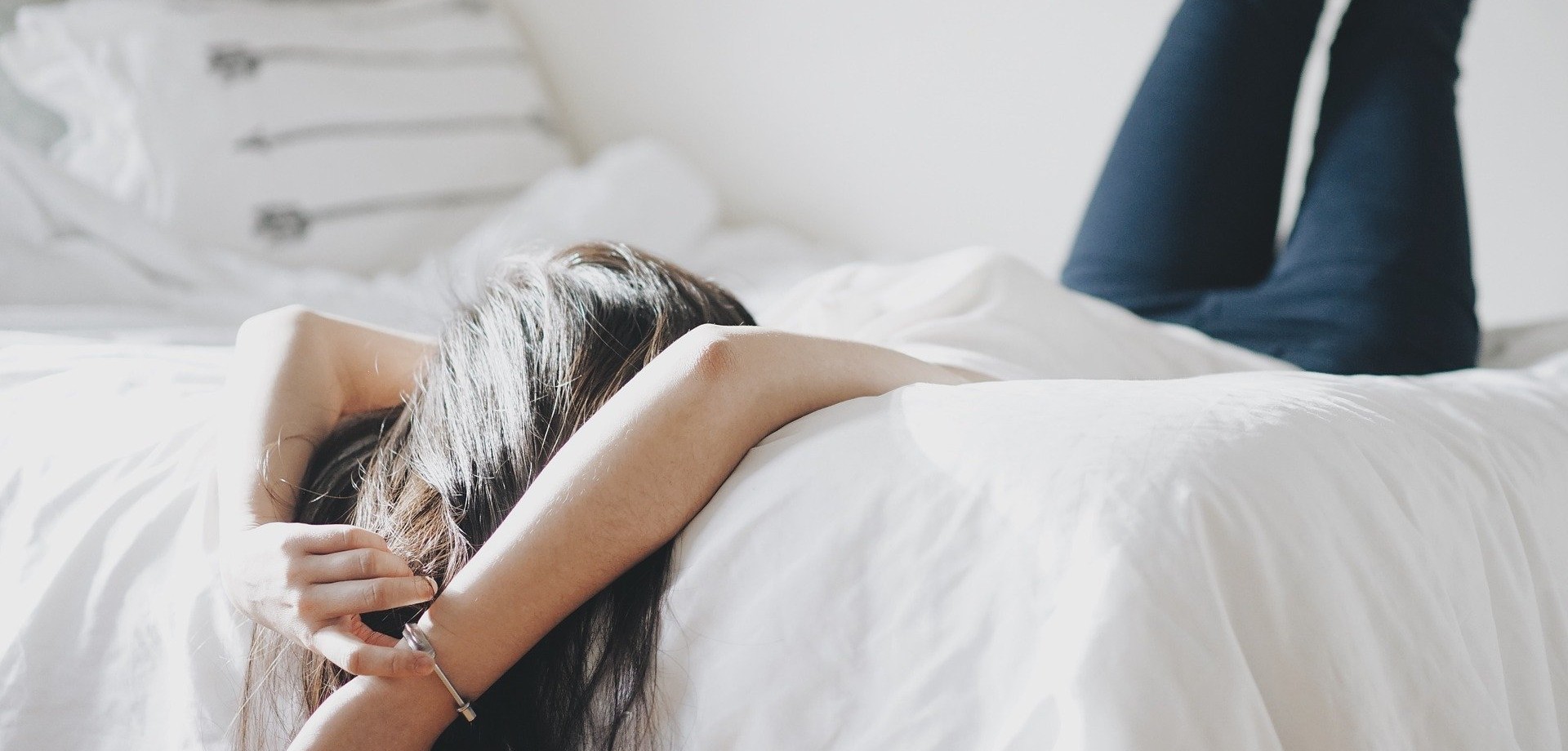 Eine junge Frau liegt auf dem Rücken in einem Bett, Arme und die langen Haare baumeln über die Matratze, die Füße sind an der Wand angelehnt