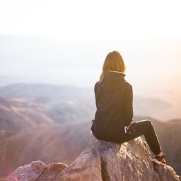 Eine junge Frau sitzt oben auf einem Felsvorsprung und blickt hinunter in das sich weit öffnende Tal